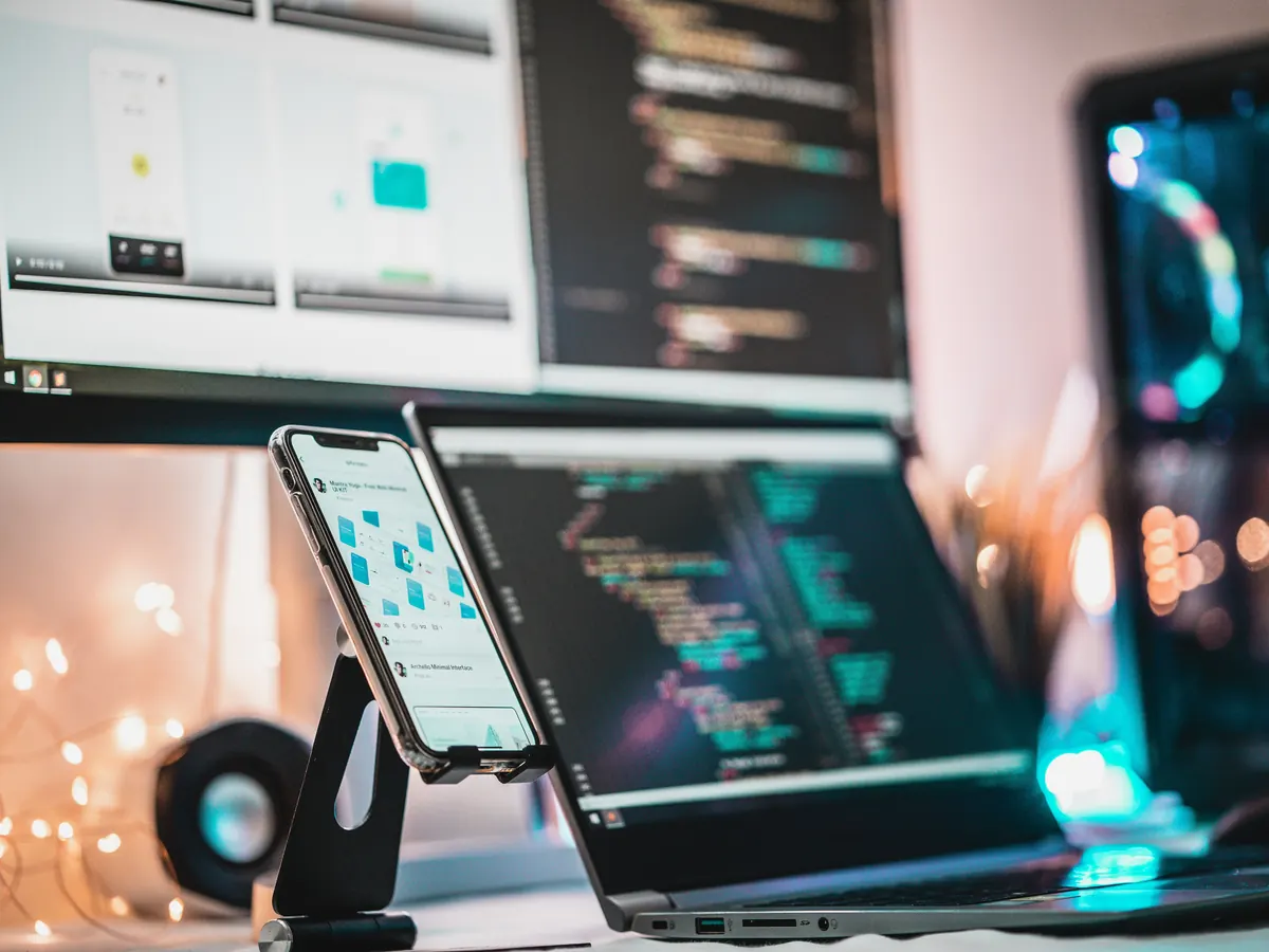Smartphone on a stand running an app beside a laptop with code and a monitor showing app screen layouts