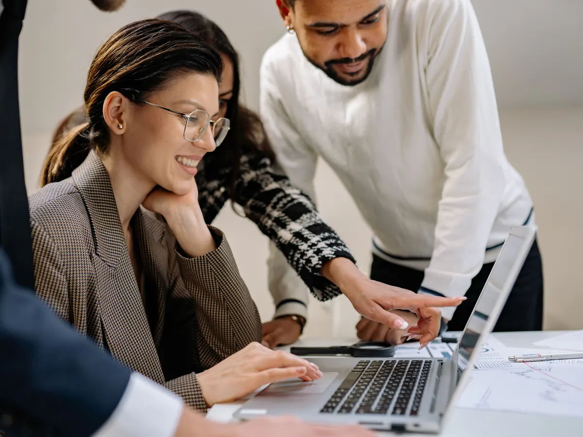 Colleagues gathered around a laptop at a shared desk, one pointing at the screen as they walk through a process together