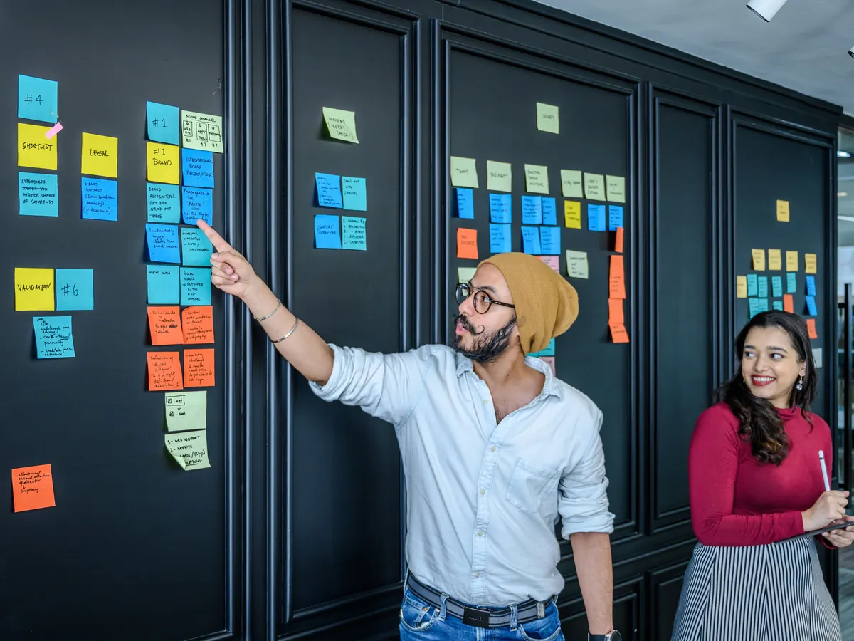 Team member pointing to sticky notes on a planning wall as a colleague looks on