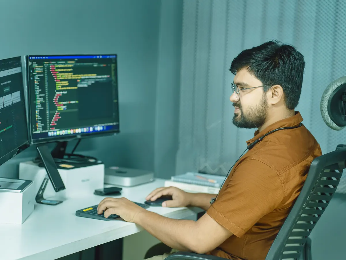 Developer in an orange shirt working through code on two monitors at a quiet, teal-toned desk