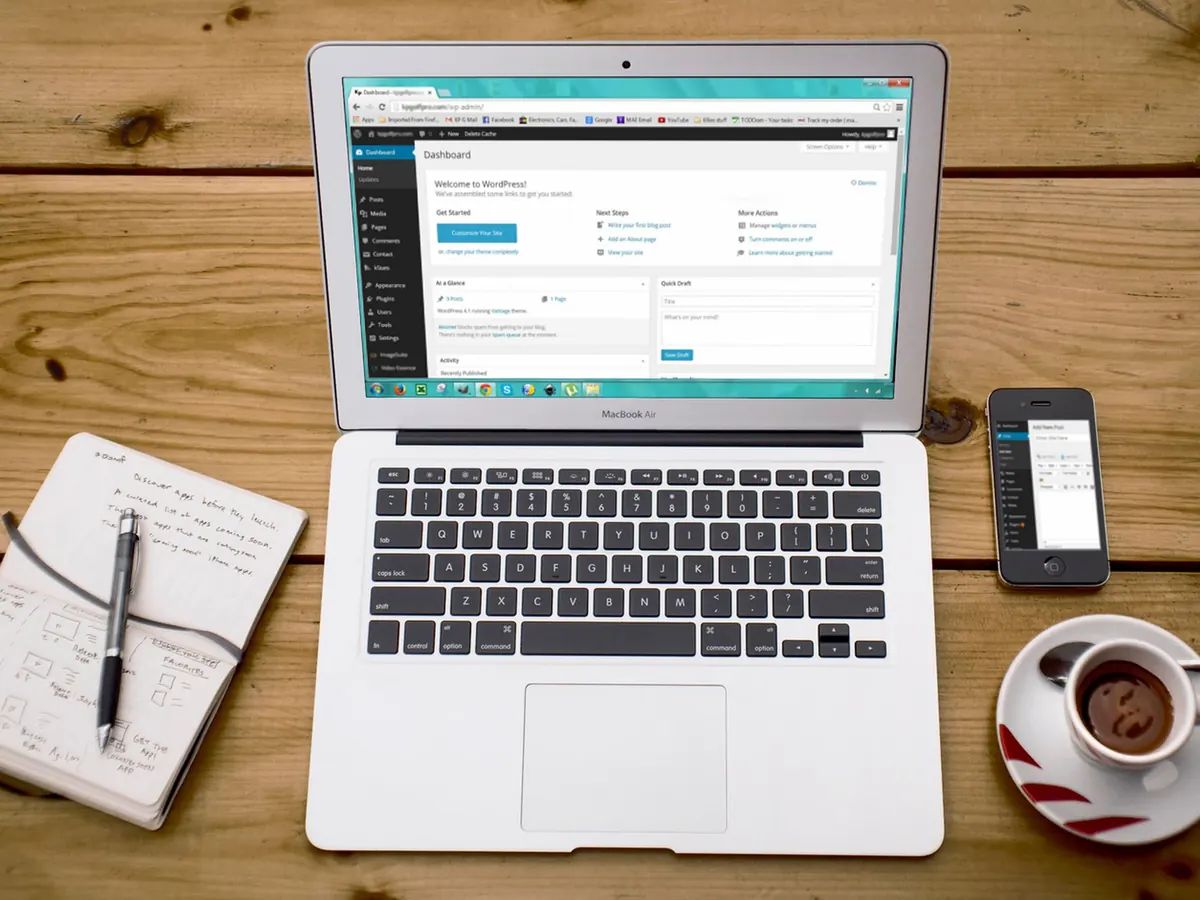 Laptop showing the WordPress admin dashboard during a WordPress development project, on a wooden desk beside a notebook, phone and coffee